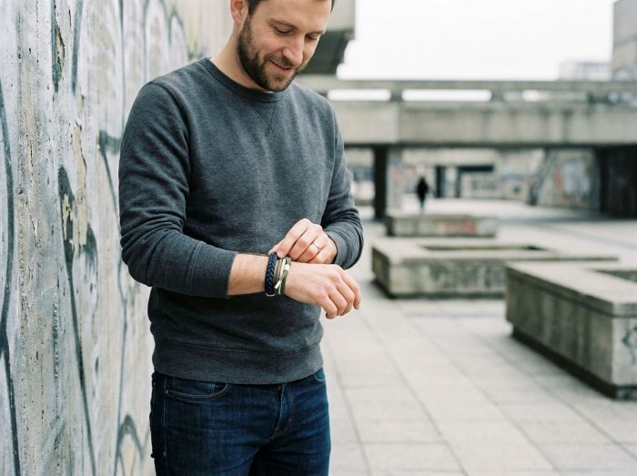 Homme en sweat gris avec un bracelet cordon marine et kaki au poignet devant un mur en béton urbain